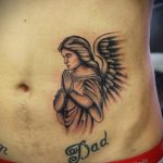 Фото тату молящегося ангела на животе. Нежный рисунок в черно-серых тонах с добавлением надписи "Dad".