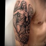 Фото татуировки ангела с розой на плече. Черно-серый рисунок с акцентом на детализацию лица и лепестков розы.