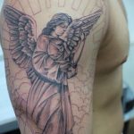 Фото тату ангела, склонившего голову в молитве на руке. Татуировка выполнена в черно-серых тонах с использованием тонких линий.
