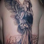 Фото тату: Черно-серый рисунок ангела с длинными волосами и крыльями на боку, на фоне мрачного пейзажа.