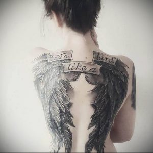 Фото тату на спине с изображением больших крыльев и цитаты "Free like a bird". Черно-белый рисунок с детально проработанными перьями и каллиграфическим шрифтом.