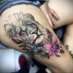 Реалистичная татуировка головы льва, окруженной розовыми и голубыми цветами, выполнена на бедре. Детализированный рисунок татуировки.
