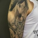 Фото татуировки на плече: реалистичное изображение лошади и тигренка, выполненное в черно-серых тонах с мягкими переходами и детальной проработкой шерсти.