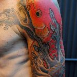 Фото татуировки с изображением красного карпа кои на плече. Рисунок выполнен в традиционном японском стиле с использованием волн и цветов.