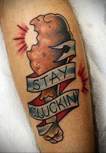 Фото татуировки с изображением куриной грудки и надписью "Stay Cluckin'" на ленте. Яркий и юмористический рисунок.