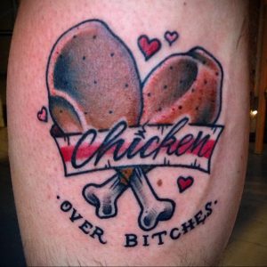 Фото тату двух куриных ножек с надписью 'Chicken Over Bitches' на руке. Яркий рисунок татуировки в стиле олдскул с использованием красных, коричневых и черных цветов.