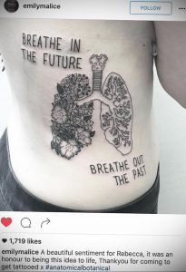 Фото тату: Большая татуировка легких, украшенных цветами, с надписью "Breathe in the future, Breathe out the past" на женском боку.