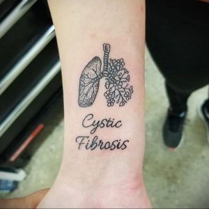 Фото тату: черная татуировка на запястье, изображающая легкие и надпись "Cystic Fibrosis". Татуировка, символизирующая борьбу с болезнью.