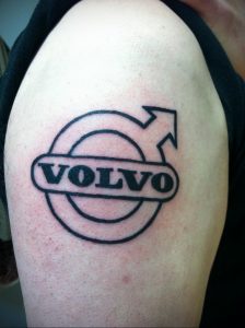 Фото тату с логотипом Volvo, выполненным в черном цвете на плече. Простой и лаконичный рисунок татуировки с изображением стрелки, направленной вверх.