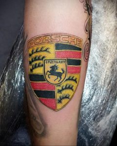 Фото тату с логотипом Porsche, выполненным в ярких цветах (красный, желтый, черный) на предплечье. Детализированный рисунок татуировки с изображением щита и надписью 'Porsche'.