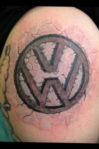 Фото тату с изображением логотипа Volkswagen на плече, как будто вырывающегося сквозь потрескавшуюся кожу. Черно-серые тона, реалистичный эффект, 3D дизайн.