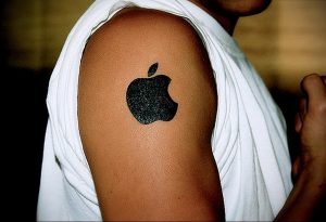 Фото тату с изображением логотипа Apple на плече. Черный силуэт надкушенного яблока, простой и лаконичный дизайн, минимализм.