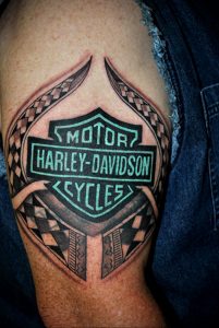 Фото тату на руке с логотипом Harley-Davidson, окруженным узорами в стиле трайбл. Черно-серые тона, детализированный рисунок, брутальный дизайн.