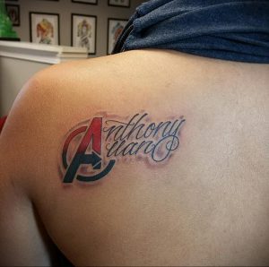 Фото тату на плече с именем 'Anthony Haro' и большой буквой 'A'. Красные и синие цвета, каллиграфический шрифт, стильный дизайн.