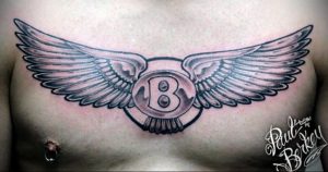 Черно-серое фото татуировки на груди с изображением буквы 'B' в круге, окруженной расправленными крыльями. Детализированный рисунок, выполненный в реалистичном стиле.