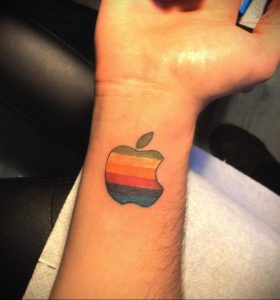 Фото тату: маленькая, цветная татуировка логотипа Apple с радужным заполнением на запястье. Изящный рисунок татуировки.