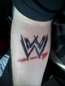 Фото тату: логотип WWE в черном цвете с красными брызгами, имитирующими кровь. Рисунок татуировки на бицепсе.