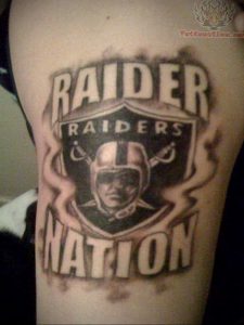 Фото тату: Логотип и надпись "Raider Nation" с изображением футбольного шлема на плече. Детализированный рисунок татуировки.
