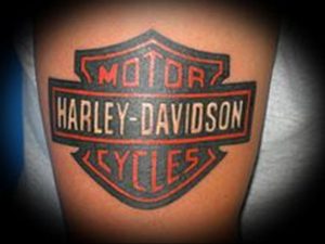 Фото тату логотипа Harley-Davidson в классическом щитовидном дизайне. Цветной рисунок татуировки, расположен на предплечье.