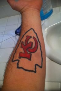 Цветная татуировка на руке с изображением логотипа команды Kansas City Chiefs в виде стрелы с буквой 'KC'.