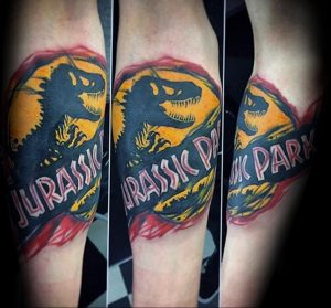 Цветная татуировка на руке с изображением логотипа фильма Jurassic Park и силуэта динозавра, выполненная в ярком стиле.