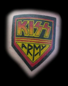 Цветная татуировка на плече с изображением логотипа рок-группы KISS в виде щита, выполненная в стиле олдскул.