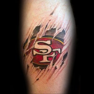 Цветная татуировка на руке с изображением логотипа команды San Francisco 49ers, как будто вырывающегося сквозь кожу.