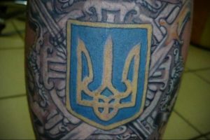 Фото тату с изображением герба Украины на руке. Сине-желтой щит с трезубцем, окруженный сложным узором.