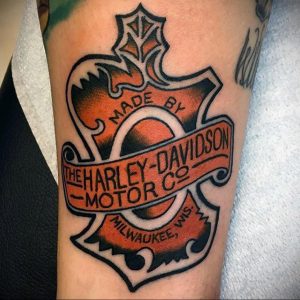 Фото тату логотипа Harley-Davidson на предплечье. Черно-оранжевый рисунок, классический дизайн, детализированная прорисовка. Подходит для поклонников мотоциклов.