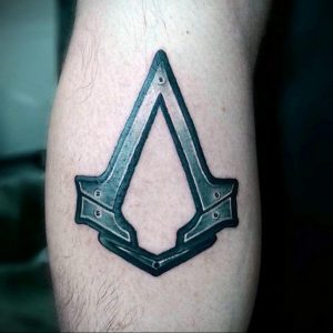 Фото тату символа Assassin's Creed на икре. Черно-серый рисунок, детализированная прорисовка, металлический блеск. Подходит для фанатов серии игр.