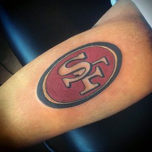 Фото тату на руке: логотип команды San Francisco 49ers в овальной рамке. Красный и золотой цвета, простой дизайн.