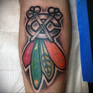 Фото тату на ноге: логотип хоккейной команды Chicago Blackhawks с тотемом и перьями. Красный, зеленый, оранжевый, серый цвета, детализированный рисунок.