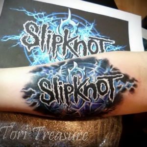 Фото тату на предплечье с изображением логотипа группы Slipknot. Черные, белые и синие цвета, динамичный стиль.