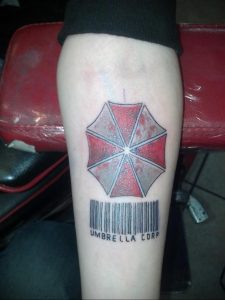 Фото тату на ноге с изображением красного зонта Umbrella Corporation из Resident Evil и штрих-кодом. Рисунок татуировки в реалистичном стиле.
