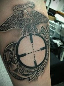 Фото тату на руке с изображением эмблемы Корпуса морской пехоты США (USMC) и прицела. Детализированный рисунок татуировки в черно-белом стиле.