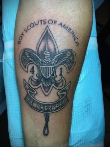 Фото тату на руке с изображением значка Boy Scouts of America. Детализированный рисунок татуировки в черно-белом стиле.