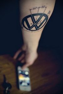 Черно-белое фото татуировки с логотипом Volkswagen на предплечье. Минималистичный рисунок татуировки, выполненный тонкими линиями.