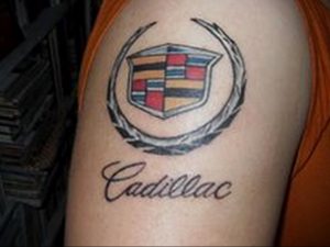 Фото тату на плече с изображением логотипа Cadillac в лавровом венке и надписью 'Cadillac'. Классический рисунок татуировки, выполненный в мягких тонах.