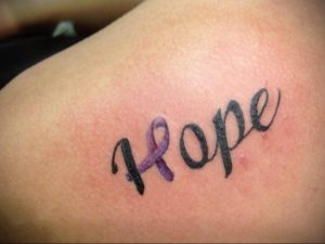 Фото тату фиолетовой ленты и надписи 'Hope' на плече. Символ надежды и поддержки, выполненный в нежной цветовой гамме.