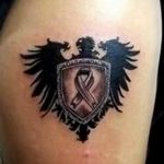 Фото тату: геральдический щит с изображением ворона и ленты, символ силы, защиты и поддержки.