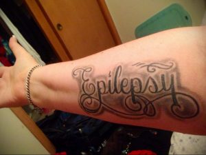 Фото тату: надпись 'Epilepsy' в обрамлении декоративных завитков на руке, символ поддержки и осведомленности.