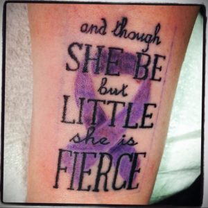 Фото тату: надпись 'and though she be but little she is fierce' с фиолетовой лентой, символ силы и мужества.