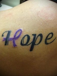 Фото тату на плече: надпись "Hope" выполнена черными и фиолетовыми чернилами в каллиграфическом стиле. Простой, но значимый рисунок, символизирующий надежду.