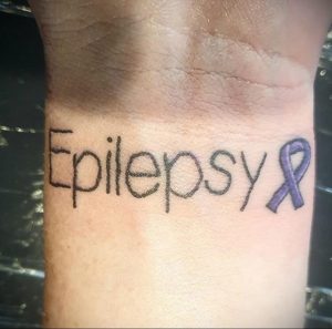 Фото тату надписи 'Epilepsy' на запястье с фиолетовой лентой. Минималистичный и значимый рисунок.