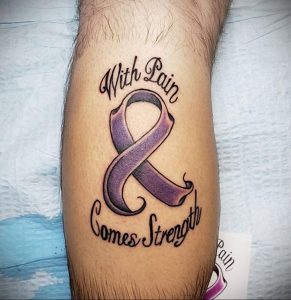 Фото тату: фиолетовая лента с надписью 'With Pain Comes Strength' на ноге. Символ стойкости и надежды, выполненный в ярких цветах.