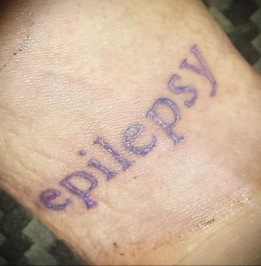 Фото тату надписи 'Epilepsy' на коже. Простой шрифт, фиолетовый цвет, минималистичный дизайн. Идея для татуировки, посвященной борьбе с эпилепсией.