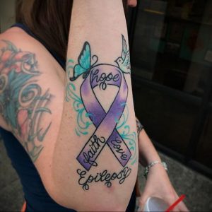 Фото тату на предплечье: фиолетовая лента, бабочка, птица и надписи 'hope', 'faith', 'love', 'Epilepsy'. Символизирует поддержку и борьбу с эпилепсией.