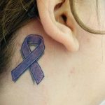 Фото тату за ухом: небольшая фиолетовая лента, символизирующая поддержку, нанесенная на кожу. Минималистичный рисунок татуировки.