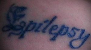 Фото тату на коже: надпись "Epilepsy" стилизованным шрифтом с небольшими крыльями, расположенная на теле. Простой рисунок татуировки.