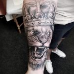 Фото татуировки на предплечье: реалистичный лев с короной, выполненный в черно-серых тонах. Детализированный рисунок символизирует силу и власть.